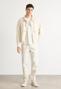 Man wearing light beige jacket, white Calvin Klein logo t-shirt, cream pants, and beige sneakers standing against a plain white background.
