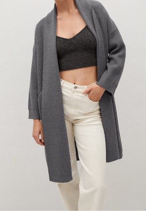 Grijze open cardigan van ribgebreid materiaal, met brede mouwen en een relaxte pasvorm, gedragen over een zwarte cropped top en witte jeans.