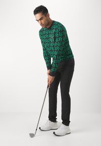 Maglione verde con motivo di logo nero ripetuto, pantaloni neri e scarpe da ginnastica bianche; golfista che si prepara a puttare con la mazza da golf in mano.