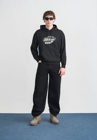 Μαύρο φούτερ με κουκούλα με το λογότυπο "Dickies", σε συνδυασμό με άνετο μαύρο παντελόνι και καφέ μποτάκια. Το μοντέλο φορά γυαλιά ηλίου, ποζάροντας μπροστά σε απλό φόντο.