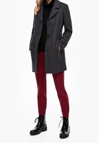 Donna che indossa un cappotto grigio scuro con bottoni, un dolcevita nero, pantaloni bordeaux e stivaletti neri lucidi stringati, in piedi con le mani in tasca.