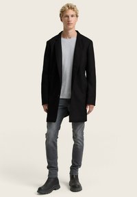 Schwarzer langer Mantel mit lockerer Passform, strukturiertem Stoff, Ein-Knopf-Verschluss, kombiniert mit grauen Jeans und schwarzen Stiefeletten.