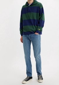 Sudadera de rayas en azul marino y verde con bolsillo tipo canguro, cuello abotonado, combinada con jeans celestes y zapatillas de lona negras.