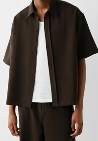 Kortärmad skjorta med knappar i mörkbrunt tyg, med krage, rak skärning och subtil glans. Bärs över ett vitt linne.