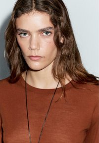 Brown, fitted top with a crew neckline. Long, wavy hair frames the face, highlighting clear skin and prominent cheekbones.