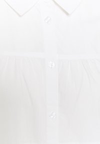 Gros plan sur le devant d'une chemise blanche à col avec des boutons translucides et un détail de tissu froncé sous la poitrine.
