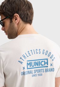 Camiseta de color beige claro con texto gráfico azul que presenta "BIENES DE ATLETISMO," "MÚNICH," y "MARCA DEPORTIVA ORIGINAL" en la parte posterior. Mangas cortas.