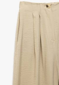 Pantaloni beige plissettati con tessuto testurizzato, dotati di chiusura a bottone visibile e tasche laterali. Design semplice e neutro.