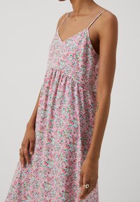 Žena v bezramenných růžových květovaných šatech s tenkými pásky, která odhaluje horní část trupu a paže s malým tetováním a prsteny na prstech.