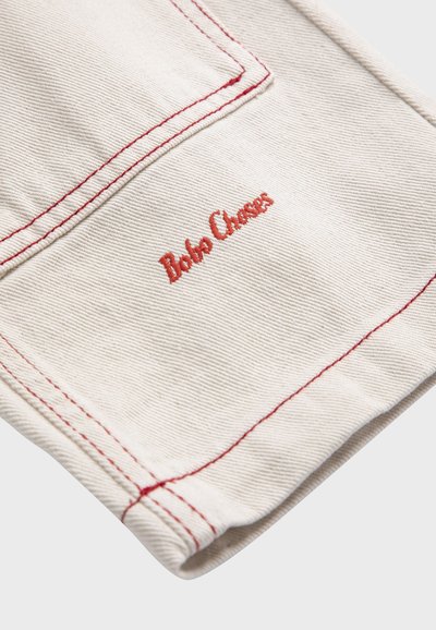 Gros plan sur une poche en tissu de couleur crème avec des coutures rouges et un texte brodé en rouge "Bobo Choses" sur un matériau en coton texturé.