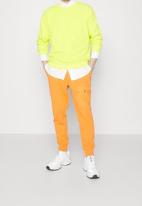 Neonově zelená mikina s žebrovanými manžetami, vrstvená přes bílou košili. Oranžové tepláky mají boční kapsu. Bílé tenisky dotvářejí outfit.