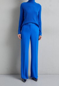 Pull à col roulé côtelé bleu associé à un pantalon large bleu. Le tissu semble lisse et légèrement brillant ; les chaussures sont foncées avec des bouts pointus.