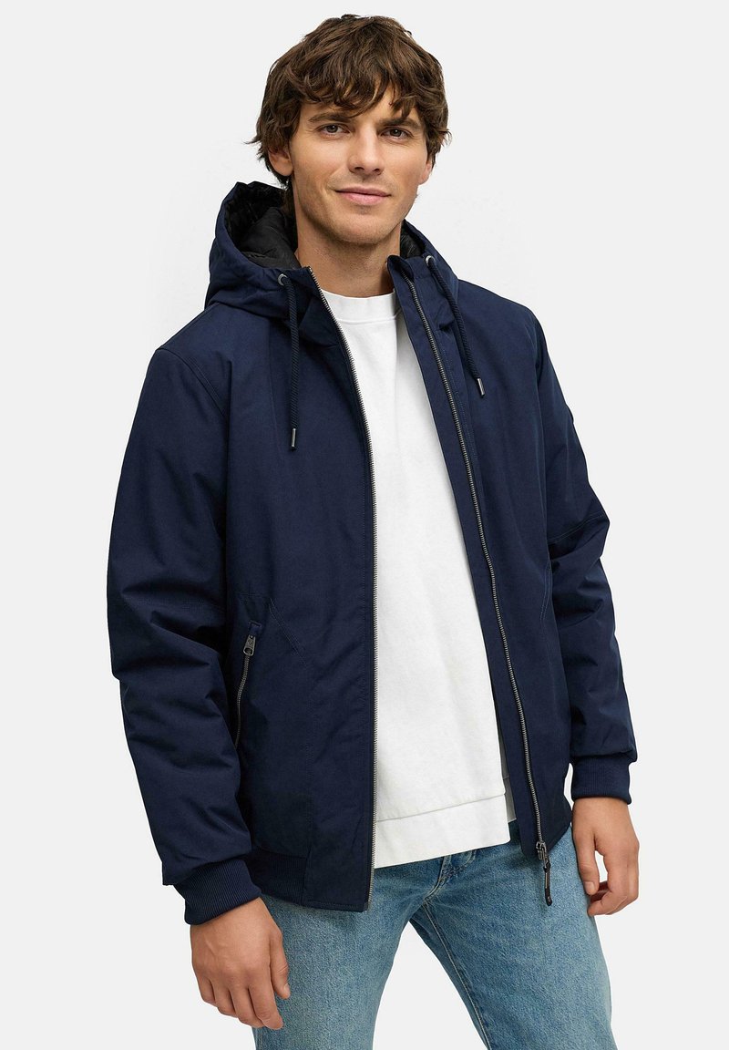 Marine vindjakke med hette, frontglidelås og sidelommer. Mansjettene og kanten har ribbestrikkede detaljer; båret over en hvit skjorte.