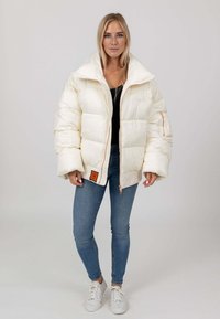 Witte pufferjack met een hoge kraag, voorzien van een rits aan de voorkant en een oranje label. Gecombineerd met lichtblauwe spijkerbroeken en witte sneakers.