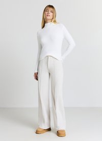 Weißer, langärmeliger Rollkragenpullover, kombiniert mit hoch taillierten, weit geschnittenen, gerippten cremefarbenen Hosen und braunen Plateau-Stiefeln, stehend vor einem hellen Hintergrund.