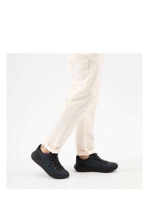 Persoon die crèmekleurige broek draagt en zwarte veterschoenen met dikke zolen, lopend op een witte achtergrond.