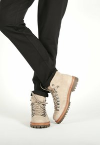 Persona con botas de senderismo de ante beige y suelas gruesas marrones, vestida con pantalones negros, de pie con las piernas cruzadas sobre un fondo blanco.