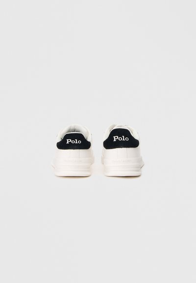 Ténis brancos com superfície texturizada, detalhes em camurça preta no calcanhar e marca "Polo" branca no calcanhar. Sola de borracha com acabamento liso.