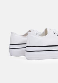 Sneaker bianche in tela con profilo basso, caratterizzate da dettagli a strisce nere sulla suola e sugli occhielli per i lacci. Texture morbida e design classico.