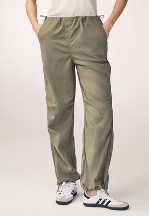 Light olive green casual pants with elastic waist and ankle drawstrings, worn with white sneakers featuring black stripes. Hands in pockets.