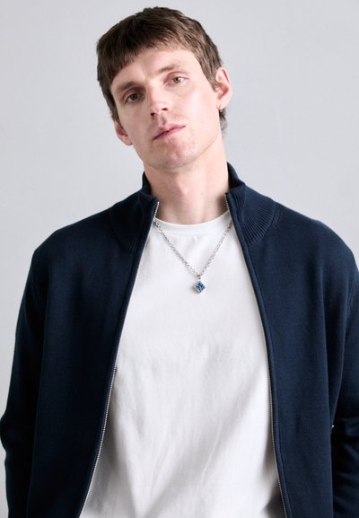 Mornarska zip-up jopica z rebrastim ovratnikom čez belo srajco. Ima srebrno verigo z modrim gem pendantom. Enobarvna ozadje.