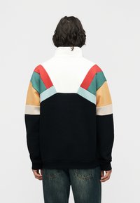 Άτομο που φοράει φούτερ με ψηλό γιακά και σχεδιασμό με χρώματα σε λευκό, κόκκινο, τιρκουάζ, μπεζ και μαύρο, απεικονιζόμενο από πίσω σε λευκό φόντο.