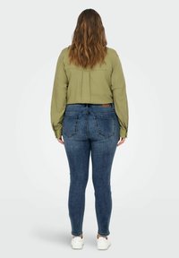 Camisa verde oliva de manga larga con pliegue en la espalda, combinada con jeans oscuros ajustados y zapatillas blancas. Vista trasera del conjunto exhibido.