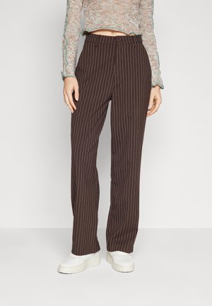 Vrouw draagt bruine pinstripe rechte broek, cropped meerkleurige gebreide trui en witte platformsneakers, staand tegen een effen achtergrond.