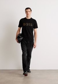 Juodas medvilninis marškinėlis su ryškiu „BOSS“ logotipu metaliniais atspalviais. Su tamsiai pilkomis džinsomis ir juodais batais, modelis laiko juodą maišą.