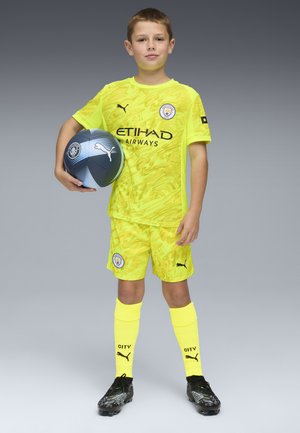 Equipación de fútbol amarilla con diseño abstracto, que presenta marcas y logotipos en negro. Botas negras y balón de fútbol azul y gris sostenido en la mano derecha.