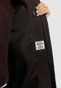 Melna quiltota jaka iekšpuse ar iešūtu kabatu un etiķeti, kas saka "Silts apskāviens no auduma." Gluda tekstūra un vienkāršs dizains.