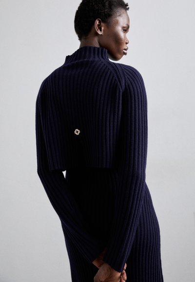 Marineblauwe ribgebreide jurk met een hoge kraag en een cropped top gedeelte. Heeft een kleine bloemaccent aan de achterkant. Lange mouwen van textuurstof.
