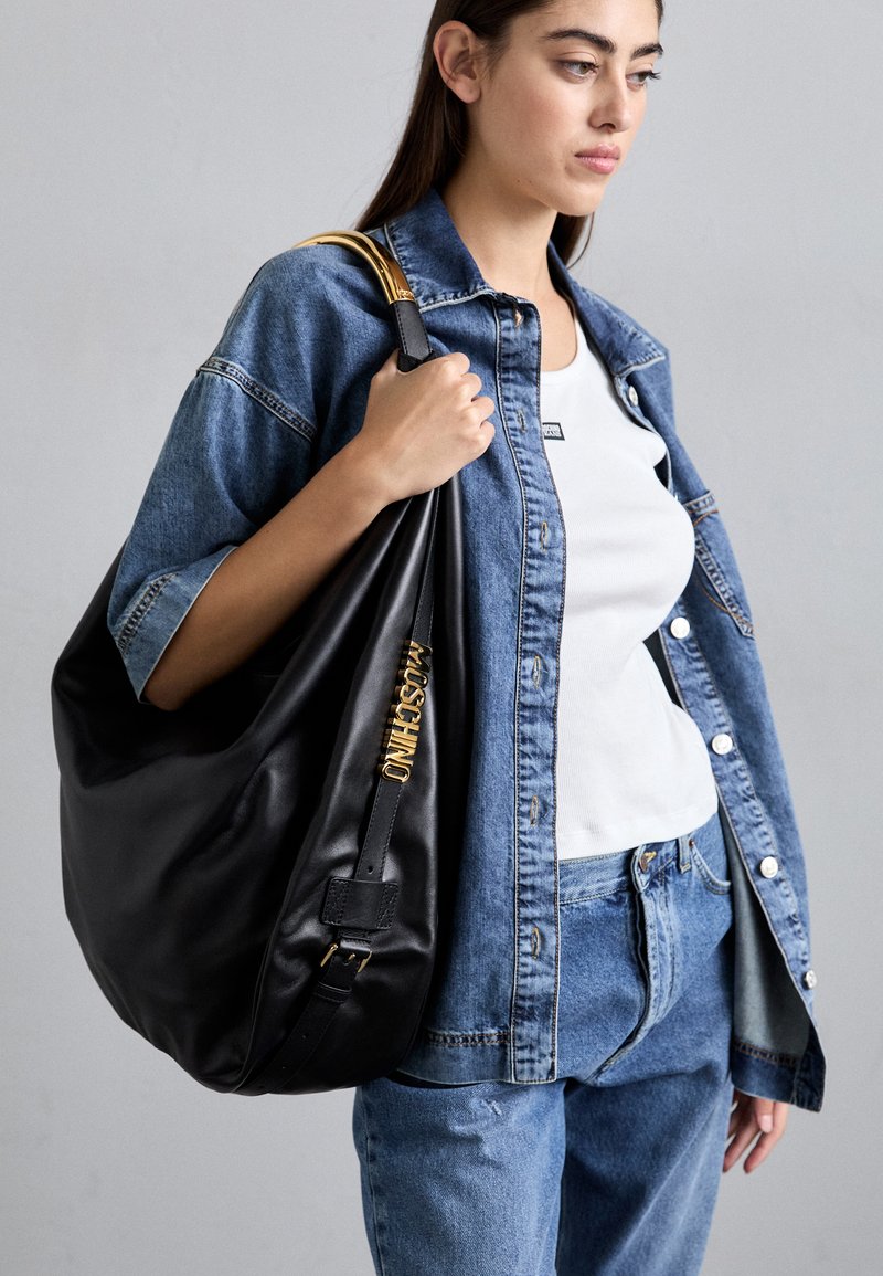 Jovem mulher vestindo uma jaqueta de jeans e calças de jeans segura uma grande bolsa de couro preta da Moschino com detalhes em dourado sobre o ombro.