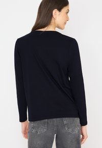 Laadukkaasta kankaaasta valmistettu tummansininen pitkähihaisuus, jossa on pyöreä kaula-aukko. Pehmeä rakenne, istuva muotoilu ja minimalistinen suunnittelu, yhdistettynä harmaisiin farkkuihin.