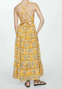 Abito maxi giallo a fiori con gonna a balze, spalline incrociate sulla schiena e motivi intricati verdi e arancioni. Arricchito da laccetti regolabili.