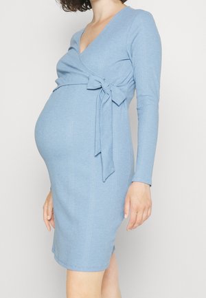 Schwangere Person, die ein hellblaues, langärmliges, geripptes Strickwickelkleid mit einer seitlich gebundenen Schleife trägt und vor einem einfarbigen Hintergrund steht.