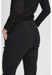 Black tailored trousers with a smooth texture, featuring back pockets and a fitted waistband. Complemented by a black, patterned lace top.