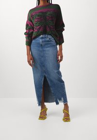 Strickpullover mit grünen und rosafarbenen Mustern, kombiniert mit einem blauen Denim-Maxi-Rock mit Schlitz vorne und ausgefranstem Saum, ergänzt durch gelbe Riemchen-High Heels.