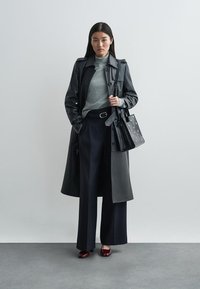 Manteau trench noir en cuir sur un col roulé gris, associé à un pantalon large navy et des chaussures rouge foncé. Sac à main noir avec des accents perforés.