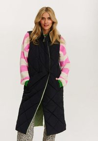 Schwarw gestepptes, langes Weste mit grüner Innenseite, ausgestattet mit einem hohen Kragen und Seitentaschen, getragen über einem gestreiften rosa- und cremefarbenen Pullover mit grünen Akzenten.