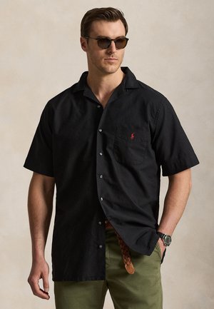 Mand iført sort kortærmet skjorte med rød logo, grønne bukser, flettet brunt bælte, ur og runde solbriller, står med hånden i lommen.