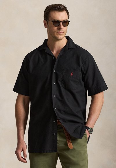 Man wearing black short-sleeve button-up shirt with red logo, green pants, braided brown belt, watch, and round sunglasses, standing with hand in pocket.