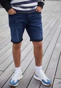 Jeansshorts i mörkblått med uppvikt fåll, tillsammans med vita och blå sneakers med orange detaljer, på en trägolv yta.