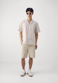 Camisa de manga corta a rayas en beige claro y blanco, combinada con pantalones cortos beige, zapatillas blancas y gafas de sol blancas.