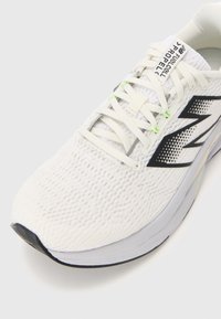 Baskets de course blanches avec une tige en mesh texturé, logo noir, accents réfléchissants et détails vert clair sur les lacets et la semelle.