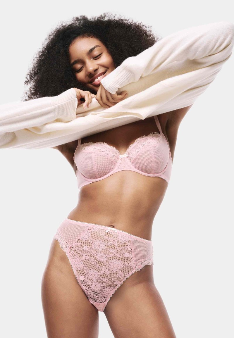 Completo di lingerie rosa chiaro con reggiseno in pizzo e slip a vita alta. I dettagli includono motivi floreali in pizzo e un accento in raso a forma di fiocco.