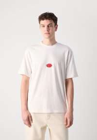 Hvid bomulds T-shirt med rund halsudskæring og korte ærmer. Har et rødt logo-patch i midten. Glat tekstur, afslappet pasform.