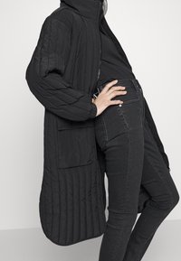 Abrigo largo acolchado negro con capucha, que presenta un cierre de cremallera y bolsillos laterales. Lleva puesto sobre jeans ajustados negros, mostrando una silueta elegante.
