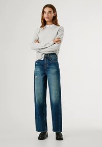 Világosszürke, kerek nyakú pulóver, magas derekú, bőszárú kék farmerrel párosítva. A farmer enyhén kifakult és kopott hatású részleteket mutat. Fekete cipő.