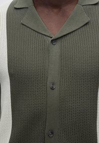 Κοντινό πλάνο ενός ατόμου που φοράει πλεκτό πουκάμισο ελιάς με υφή, γιακά, τρία κουμπιά και λευκές πλευρικές πιέτες.